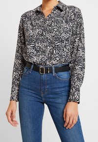 Black and white patterned long-sleeve shirt with a collar, worn with high-waisted blue jeans and a black belt featuring a gold buckle.
