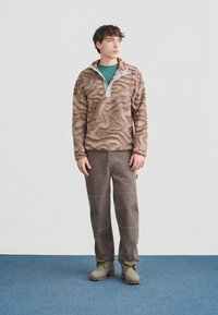 Νέος άνδρας στέκεται σε μπλε δάπεδο φορώντας καφέ fleece μπουφάν με σχέδια, πράσινο πουκάμισο, καφέ παντελόνι και μπεζ πεζοπορικά μποτάκια, κοιτάζοντας προς τα δεξιά.