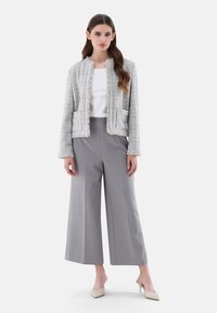 Grau melierter Blazer mit fransigen Kanten, kombiniert mit weiten grauen Hosen und nudefarbenen Heels. Darunter ein weißes Top mit minimalistischem Design.