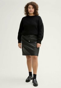 Zwarte gebreide trui met lange mouwen, gecombineerd met een aansluitende zwarte leren kokerrok. Model draagt zwarte sokken en zware loafers.