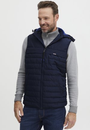 Marineblå puffervest med hette, med vertikalt quiltet design, glidelås og sidelommer. Lett stoff med en glatt tekstur.
