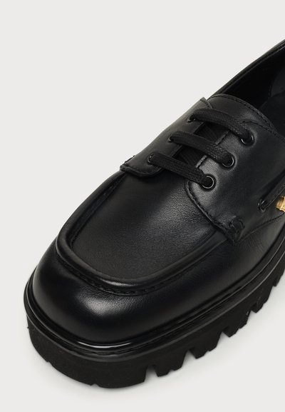 Chaussure noire en cuir à lacets avec semelle en caoutchouc épaisse et couture moc toe sur fond blanc.