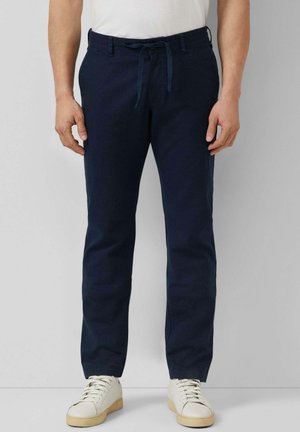 Mężczyzna w granatowych casualowych spodniach ze sznurkiem i białych sneakersach, stojący na jasnym, jednolitym tle.