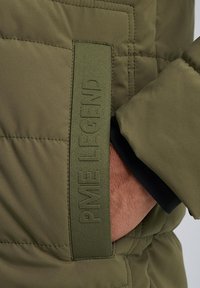 Olive green padded jacket with a textured fabric, featuring a visible brand label on the pocket and elastic cuffs for a snug fit.