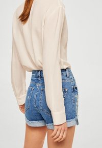 Shorts en denim bleu taille haute avec un ourlet effiloché, ornés de broderies florales sur le côté gauche et les poches arrière. Portés avec un chemisier léger.