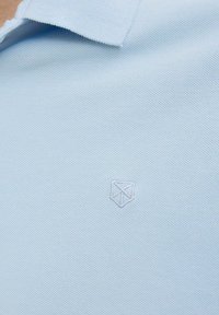 Svetlomodrá polo košeľa s textúrovanou tkaninou, obsahujúca malé biele vyšívané logo shield na ľavej strane hrude.