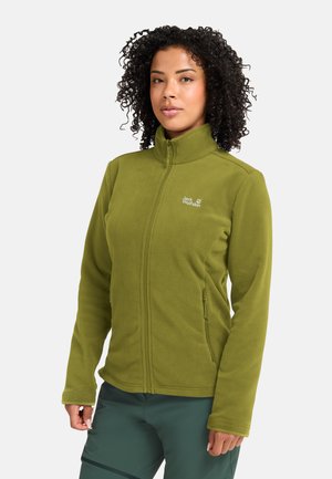 Vrouw met krullend haar die een groen fleecejack met rits en donkergroene broek draagt, staand voor een effen lichte achtergrond.