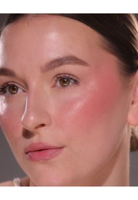 Nærbilde av et ansikt med glatt, lys hud; myk rosa rouge på kinnene; naturlig øyemakeup og glansfulle rosa lepper; rette øyenbryn med subtil bue.