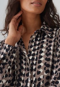 Blouse with a buttoned collar, featuring a mix of large black and white polka dots on a taupe background. Soft fabric with a smooth texture.