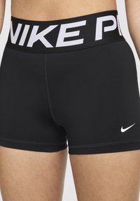 Svarte treningsshorts laget av elastisk stoff med bred linning som har "NIKE" i hvite bokstaver. Lite hvitt Nike-logo på siden.