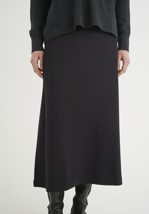 Femme portant une longue jupe noire, un pull gris foncé et des bottes noires montantes, debout devant un fond clair uni.