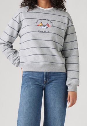 Femme portant un sweat-shirt Levi's gris clair rayé avec des drapeaux croisés et le texte "Since 1873", associé à un jean bleu.