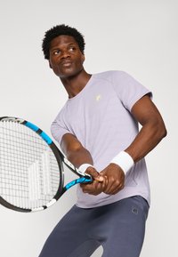 Camiseta deportiva de color púrpura claro con mangas cortas, que presenta un pequeño logo amarillo, combinada con pantalones ajustados de color azul oscuro. Racket de tenis sostenido en las manos.