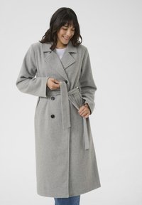 Cappotto in misto lana grigio con design doppiopetto, colletto a revers e vita legata. Presenta bottoni neri e maniche ampie.