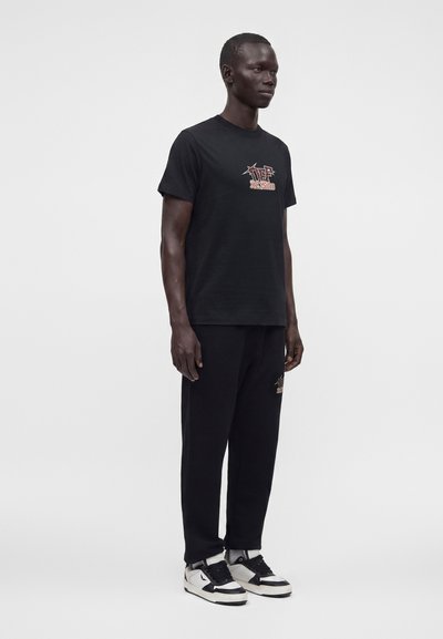 Homme portant un t-shirt graphique noir et un pantalon noir avec des baskets blanches et noires, debout devant un fond clair uni.