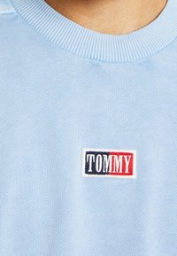 Světle modrý pletený límec trička s malou obdélníkovou nášivkou, která zobrazuje "TOMMY" v bílé, námořnické a červené barvě.