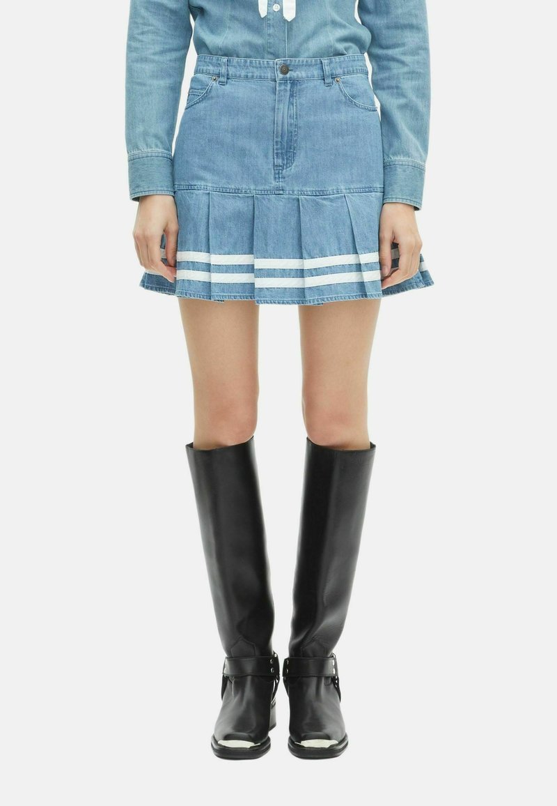 Vrouw in een blauwe denim geplooide mini rok met witte strepen en zwarte leren laarzen tot de knie, staand tegen een effen achtergrond.