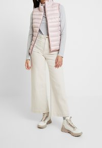 Gesteppte rosa Weste mit hohem Kragen, graues Langarmshirt, weite cremefarbene Hose und beige Schnürstiefel mit dicker Sohle.