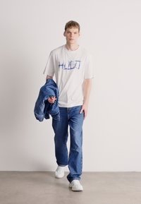 Tricou alb cu mânecă scurtă și text grafic albastru, asortat cu blugi albaștri și o jachetă cu dungi albastre. Modelul poartă adidași albi.