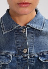 Jeansjacka i medelblått med klassisk krage, silverknappar med "Lee"-logotyp och subtila sömnadsdetaljer på fickorna.