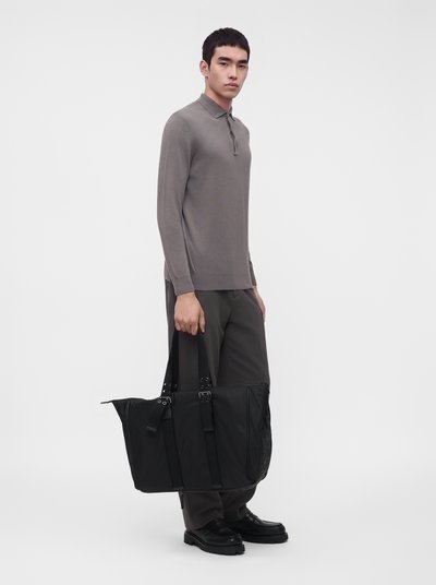 Homme portant un polo gris à manches longues et un pantalon sombre tenant un grand sac cabas noir sur un fond clair uni.