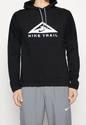 Person iført sort Nike Trail-hoodie med hvidt logo og grå Nike-bukser med hvid swoosh, stående med hænderne afslappet ned langs siden.