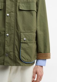 Πράσινο μπουφάν με κουμπιά πατιλέτας, μεγάλες τσέπες με καπέλο, μανσέτες από καφέ βελούδο, φορεμένο πάνω από ανοιχτό μπλε πουκάμισο και άσπρο παντελόνι, με το δεξί χέρι ορατό.