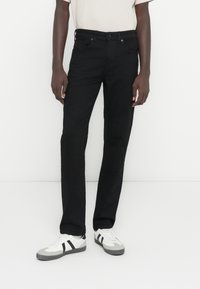 Fekete egyenes szabású farmer, ötkantoros kivitel, gombos záródással, fehér sportcipőkkel, amelyeken fekete díszítések találhatók.