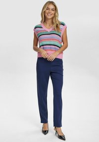 Gebreide, mouwloze v-hals trui met meerkleurige horizontale strepen in roze, groen, blauw en zwart; gecombineerd met marineblauwe op maat gemaakte broek en zwarte hakken.