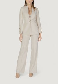 Blazer beige sartoriale con rever a sacchetto, chiusura con un solo bottone e dettagli in oro, abbinato a pantaloni a gamba larga coordinati.