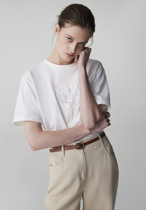 Joven con piel clara y ojos azules, vestida con una camiseta blanca con bordado de flores, pantalones beige y cinturón marrón, posando frente a un fondo liso.