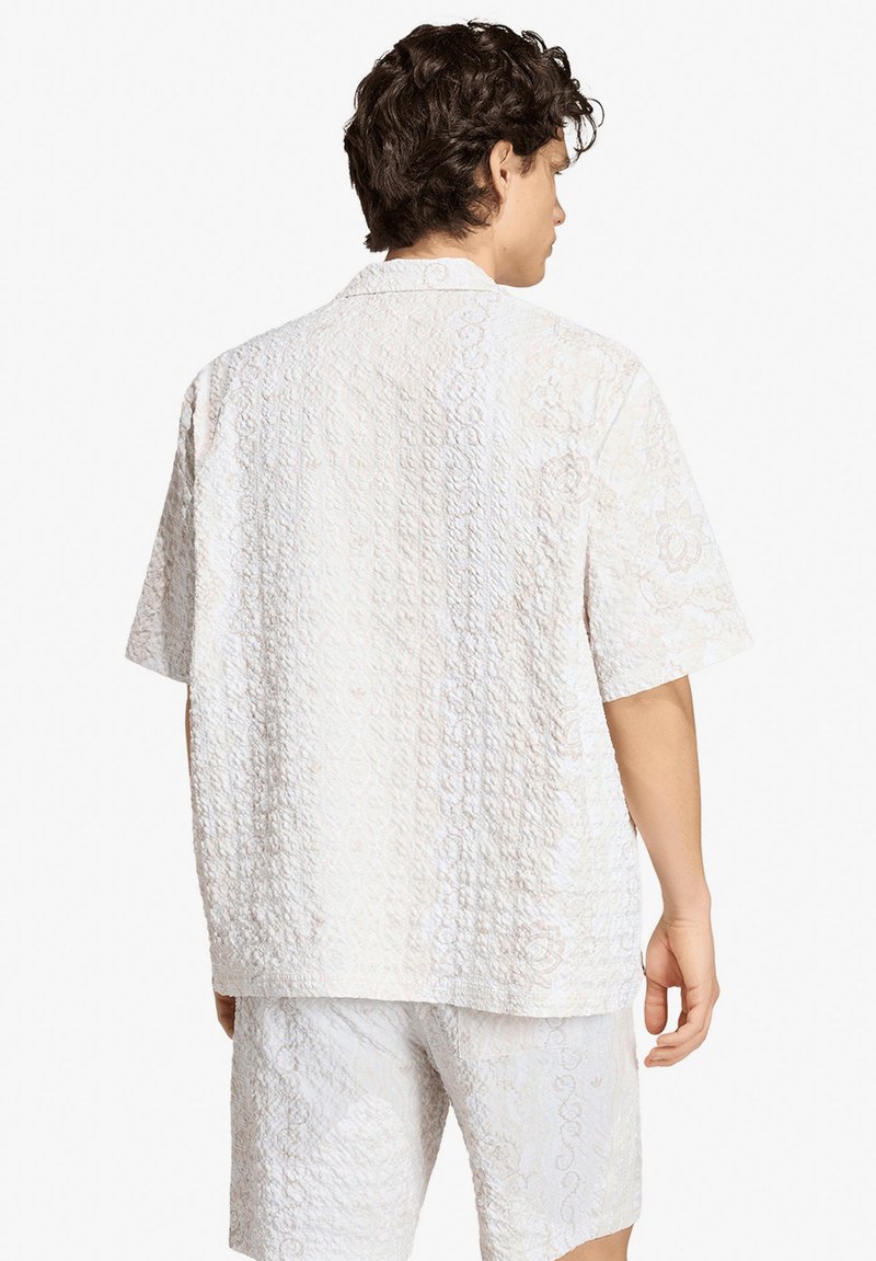 Uomo visto da dietro che indossa una camicia bianca a maniche corte texturizzata e pantaloncini abbinati con un sottile motivo floreale.