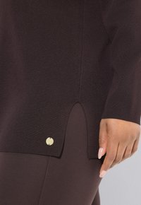 Pull en maille marron foncé avec une fente sur le côté et une petite étiquette en or ronde près de l'ourlet, porté avec un pantalon assorti et une main visible.