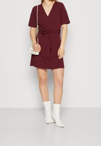 Vestido envolvente marrón con mangas cortas, cintura ceñida con cinturón y un corte relajado. Combinado con botas tobilleras blancas y un pequeño bolso blanco.