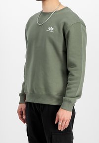 Sudadera verde oliva con puños y dobladillo acanalados, cuello redondo, con un logo blanco en el pecho y un detalle rojo en el lateral.