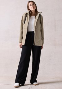 Veste imperméable beige clair avec une capuche, fermeture éclair à l'avant et cordons de serrage noirs, associée à un pantalon noir ample et des baskets blanches.