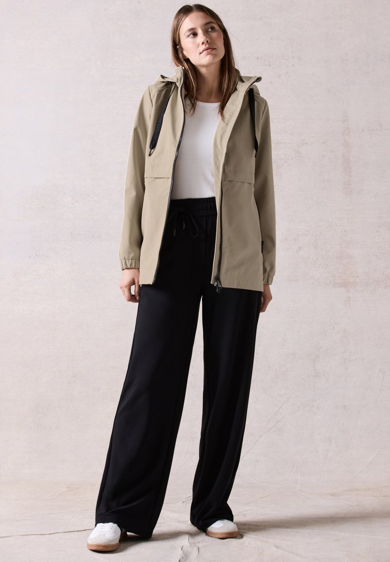 Veste imperméable beige clair avec une capuche, fermeture éclair à l'avant et cordons de serrage noirs, associée à un pantalon noir ample et des baskets blanches.