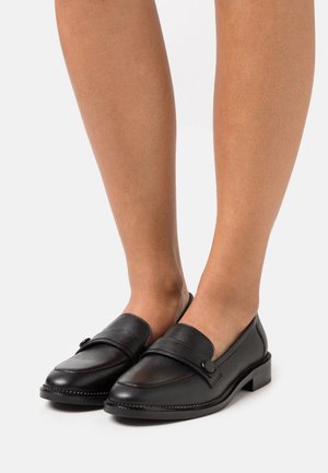 Musta nahkakengät, joissa on pyöreä varvas, koristeellinen nahkaremmi, pieni napin yksityiskohta ja matala kanta. Tasainen pinta, jossa on erottuvaommeltu yksityiskohta.