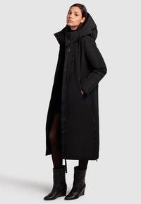 Black long coat with a hood, zip closure, multiple zippered pockets, and textured fabric. Worn over fitted black attire and ankle boots.
