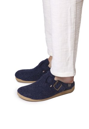 Zapatillas de casa de felpa azul marino con suela de caucho beige, que presentan una hebilla lateral y una superficie texturizada, combinadas con pantalones blancos texturizados.