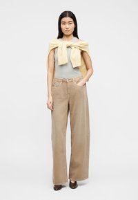 Γυναίκα που φοράει αμάνικο γκρι τοπ, μπεζ παντελόνι ψηλόμεσο με φαρδιές παντελόνες, καφέ παπούτσια, με κίτρινο πουλόβερ δεμένο στους ώμους της.