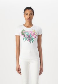 T-shirt blanc à manches courtes présentant un motif tropical vibrant en rose et vert. Le design comprend des feuilles et une forme triangulaire géométrique.