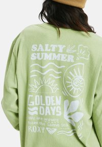 Jasnozielona bluza z białymi grafikami nadrukowanymi, w tym tekstem, słońcem, muszlą i falami na plecach. Miękki, luźny krój.