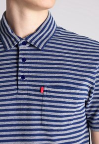 Homme portant un polo rayé horizontal bleu et blanc avec un col et des boutons bleus, sur fond clair.