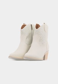 Ζευγάρι μπεζ μποτάκια αστραγάλου με υφή στην επιφάνεια, φερμουάρ στο πλάι, θηλυκές λαβές και ψηλά τακ ούνια τύπου block σε λευκό φόντο.