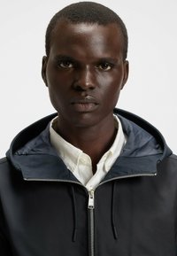 Chaqueta negra con cremallera y capucha, con textura suave, cremallera plateada y forro en capas de color marino sobre una camisa blanca con cuello.