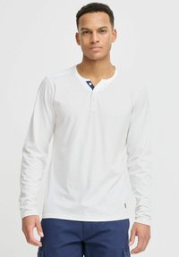 Hvit henley-skjorte med lange ermer og knappet halsåpning, med en kontrasterende marineblå krage. Stoffet ser glatt ut og viser en klassisk passform.