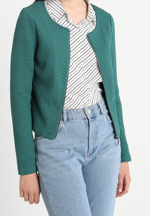 Kobieta w tealowym, teksturowanym, zapinanym na zamek kurtce, nosząca białą i czarną w paski koszulę oraz jasnoniebieskie, wysokie dżinsy, na tle jednolitego tła.