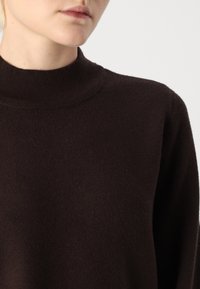Mørkebrun genser med ribbet høy hals, skreddersydd passform og strukturert stoff. Ermene er litt avslappet med en glatt overflate.