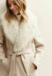 Abrigo beige con un gran y suave collar de piel sintética. Presenta un cinturón atado en la cintura y dos bolsillos laterales. Textura suave y diseño elegante.